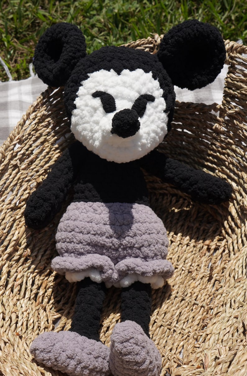 May include: A handmade crochet doll resembling a cartoon character. The doll features a black body, ears, and arms, with a white face and a black nose. It wears gray shorts and gray shoes. The doll is placed on a woven basket.