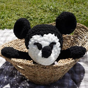 May include: A handmade black, white, and tan crochet Mickey Mouse plush toy. The plush is in a woven basket on a checkered blanket, with green grass in the background. The plush has large black ears and a white face.