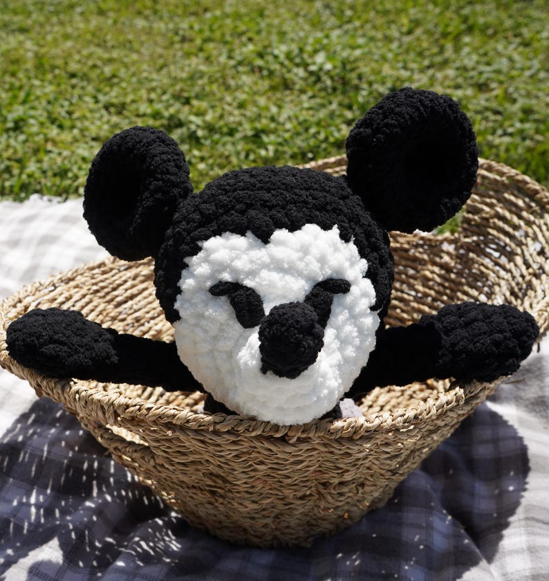 May include: A handmade black, white, and tan crochet Mickey Mouse plush toy. The plush is in a woven basket on a checkered blanket, with green grass in the background. The plush has large black ears and a white face.
