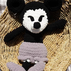 May include: A handmade, plush Mickey Mouse doll. The doll features black ears, arms, and body, with a white face and a black nose. It wears gray shorts and gray feet. The doll is sitting in a woven basket.