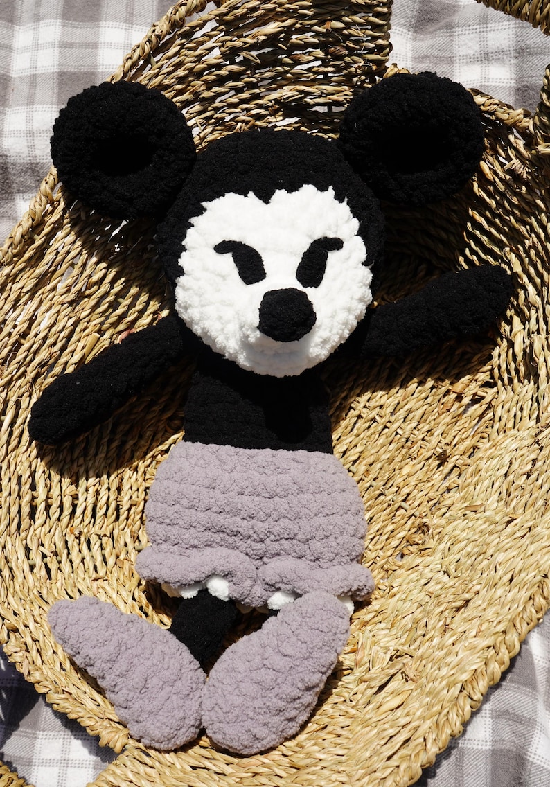 May include: A handmade, plush Mickey Mouse doll. The doll features black ears, arms, and body, with a white face and a black nose. It wears gray shorts and gray feet. The doll is sitting in a woven basket.