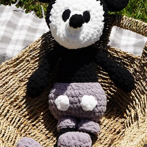 May include: A handmade crochet plush toy of a cartoon mouse character. The toy features a black body, ears, and arms, with a white face and a gray bottom. It is sitting in a woven basket on a checkered blanket.