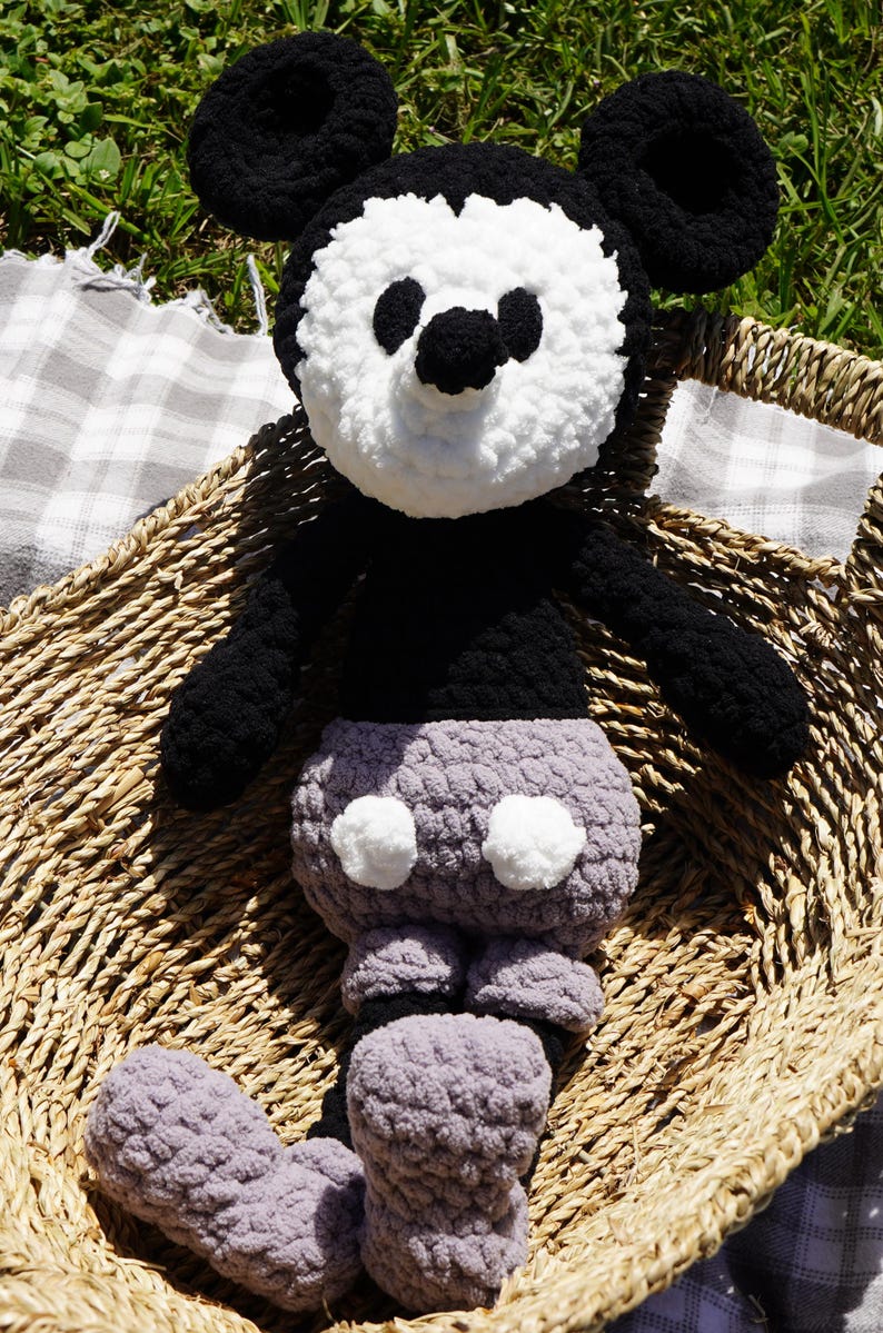 May include: A handmade crochet plush toy of a cartoon mouse character. The toy features a black body, ears, and arms, with a white face and a gray bottom. It is sitting in a woven basket on a checkered blanket.