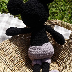 May include: A handmade crochet Mickey Mouse doll. The doll has a black head, ears, arms, and legs, with a gray body and feet. It is sitting in a woven basket, with a checkered cloth visible in the background.