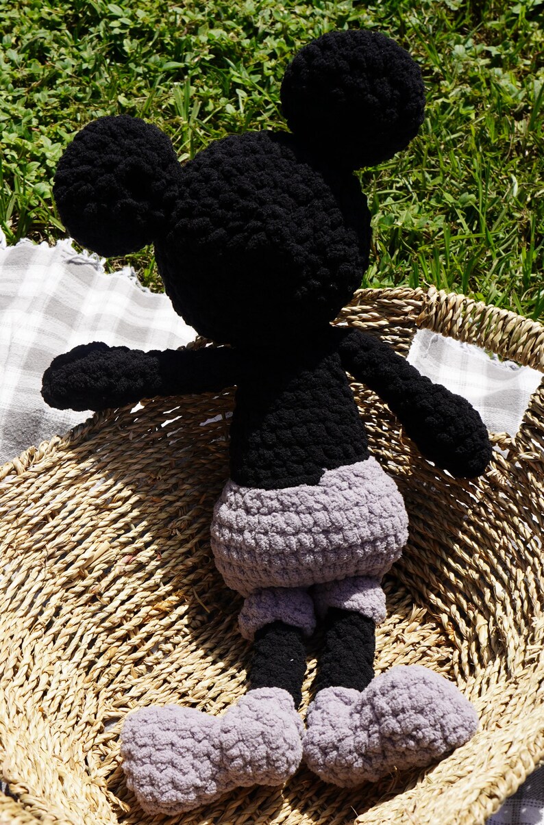 May include: A handmade crochet Mickey Mouse doll. The doll has a black head, ears, arms, and legs, with a gray body and feet. It is sitting in a woven basket, with a checkered cloth visible in the background.