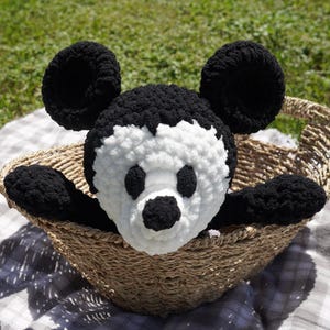 May include: A handmade black and white crochet Mickey Mouse plush toy with large round ears, black eyes, and a black nose. The plush is in a woven basket, with its arms extending over the edge. The background is a grassy area.