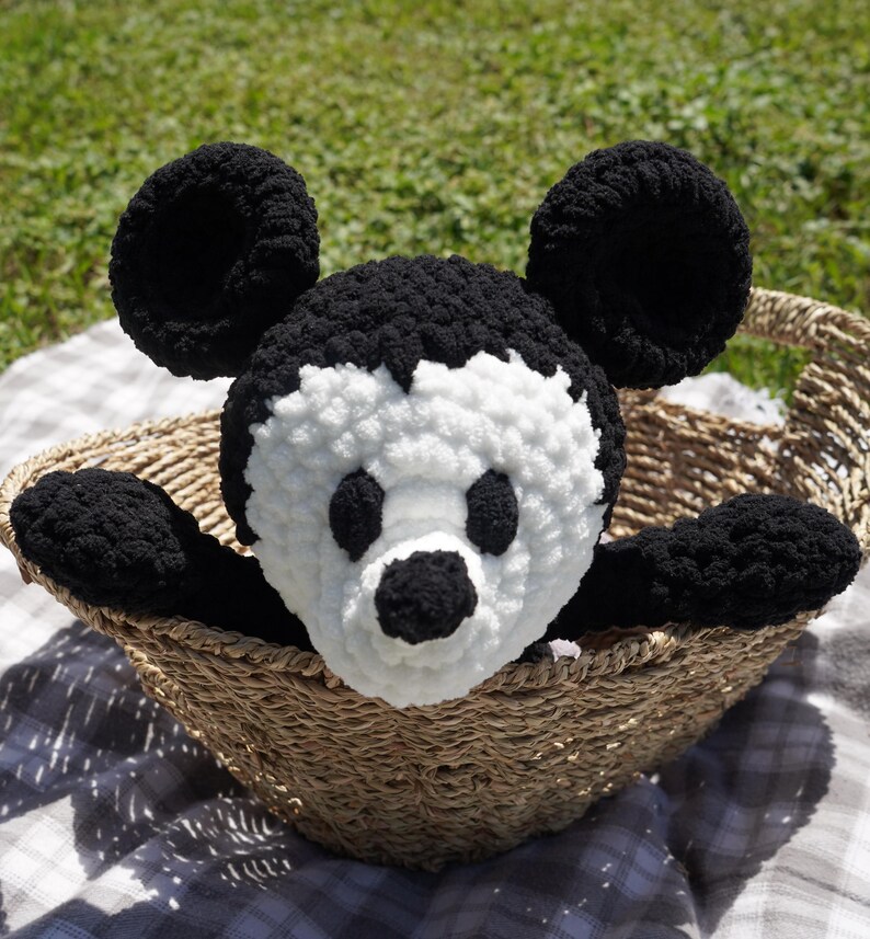 May include: A handmade black and white crochet Mickey Mouse plush toy with large round ears, black eyes, and a black nose. The plush is in a woven basket, with its arms extending over the edge. The background is a grassy area.
