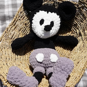 May include: A handmade crochet Mickey Mouse plush toy. The plush features black ears, arms, and feet, a white face with black eyes and nose, and gray shorts with white buttons. The toy is sitting in a woven basket.