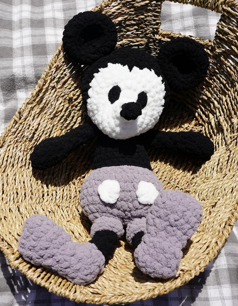 May include: A handmade crochet Mickey Mouse plush toy. The plush features black ears, arms, and feet, a white face with black eyes and nose, and gray shorts with white buttons. The toy is sitting in a woven basket.