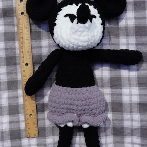 May include: A handmade crochet doll resembling a cartoon character. The doll has a black body, arms, and legs, with a white face and ears. It wears a gray skirt. The doll is placed on a gray and white checkered surface.