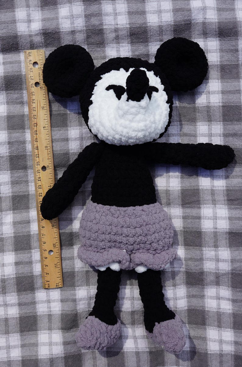 May include: A handmade crochet doll resembling a cartoon character. The doll has a black body, arms, and legs, with a white face and ears. It wears a gray skirt. The doll is placed on a gray and white checkered surface.