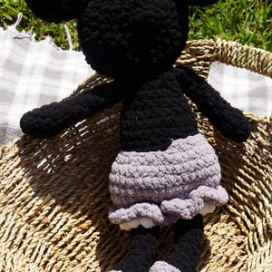May include: A handmade black and gray crochet doll with large round ears, arms, and legs. The doll has a gray skirt with a ruffled hem and is sitting in a woven basket. The doll's feet are gray and black.