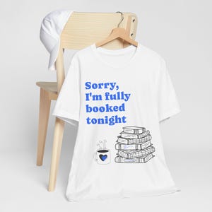Puede incluir: Camiseta blanca con el texto azul "Sorry, I'm fully booked tonight." Una pila de libros y una taza de café con un corazón están debajo del texto. Una gorra de béisbol blanca descansa sobre una silla de madera.