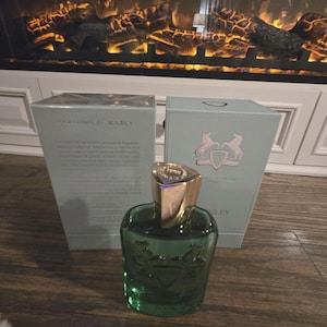 May include: A green glass perfume bottle with a gold cap sits in front of two light blue boxes. The bottle has a decorative design and the boxes have text and a crest logo. The boxes read "Parfums de Marly".