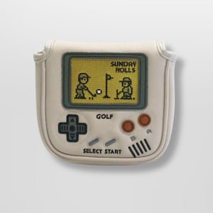 May include: A cream-colored golf club headcover with a retro video game design. The cover features a screen displaying a pixelated golf scene with the text "SUNDAY ROLLS" and "GOLF". It has buttons and a directional pad, resembling a classic handheld console.