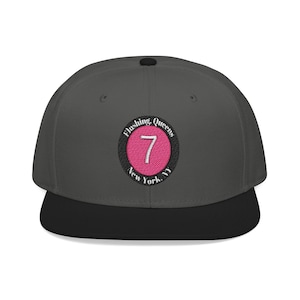 Op de afbeelding: Een antracietgrijze baseballcap met een zwarte klep. De pet heeft een geborduurde ronde patch met de tekst "Flushing, Queens, New York, NY" rond een roze cirkel met het nummer "7" in het midden.
