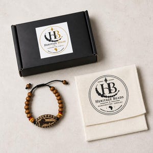 Könnte beinhalten: Ein Armband aus braunen Holzperlen mit einer zentralen ovalen Perle mit dunklem Ätzmuster sowie kleinen roten und gelben Akzentperlen. Das Armband liegt neben einer schwarzen rechteckigen Schachtel und einem cremefarbenen Stoffbeutel, die beide das Heritage Beads-Logo tragen.