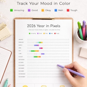 May include: A clipboard with a 2026 Year in Pixels mood tracker. The tracker uses color-coded squares to represent moods, with options like "Amazing," "Good," and "Tough." A hand is shown using a purple pen to fill in the squares. The text "Track Your Mood in Color" is displayed.