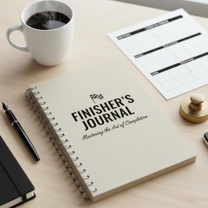 May include: A cream-colored spiral-bound notebook with the text "FINISHER'S JOURNAL" and a checkered flag graphic. A cup of coffee, a pen, and a planner are also on the light-colored wooden surface.