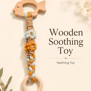 May include: A wooden fish-shaped teether attached to a braided macrame cord in tan, light blue, and mustard yellow, featuring two small flower-shaped knots and a round wooden ring at the base. The text reads Wooden Soothing Toy Teething Toy.