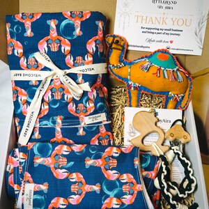 May include: A gift box containing a dark blue fabric with a lobster and bubble pattern tied with a ribbon labeled WELCOME BABY, a felt camel ornament, a wooden animal-shaped teether, and a black and white macrame pacifier clip with wooden beads.