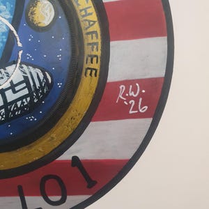K&ouml;nnte beinhalten: Rundes Gem&auml;lde mit dem Apollo 1 Missionsabzeichen. Das Design zeigt ein Raumschiff, einen Planeten und einen Sternenhimmel. Der Rand ist gelb mit dem Namen "CHAFFEE" in Schwarz. Der Hintergrund hat rote und wei&szlig;e Streifen, mit der Nummer "01" in Schwarz.