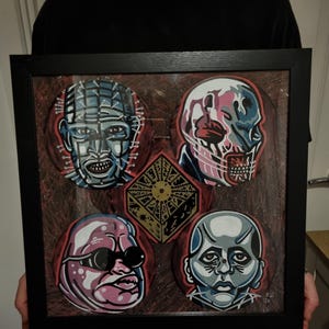 May include: A framed artwork featuring stylized portraits of characters from the Hellraiser film series. The artwork includes five circular portraits and a central geometric shape, all set against a textured background. The frame is black.