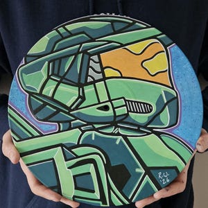 May include: A circular artwork with a green and teal helmet design, featuring an orange visor, set against a blue backdrop with white dots. The artwork includes the initials "R.W. '26".