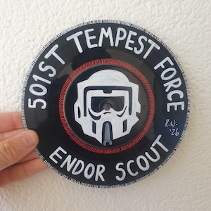Peut inclure: Un disque vinyle noir avec des lettres blanches et un dessin de casque blanc. Le texte indique "501st Tempest Force Endor Scout". Un cercle rouge entoure le casque. Signé "R.W. '26".