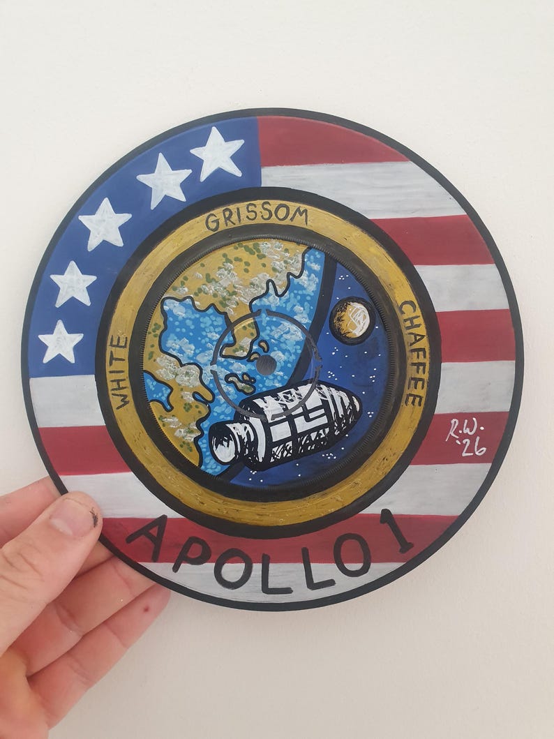 K&ouml;nnte beinhalten: Handgemalte Apollo 1 Gedenkkunst auf einer kreisf&ouml;rmigen Oberfl&auml;che. Das Design zeigt die amerikanische Flagge, Sterne und Streifen, mit den Namen "Grissom, White, Chaffee" und dem Wort "Apollo 1". Eine detaillierte Illustration eines Raumschiffs ist ebenfalls enthalten.