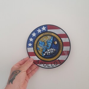 K&ouml;nnte beinhalten: Handgemalte, runde Apollo 1 Gedenktafel. Das Design zeigt ein zentrales Bild eines Raumschiffs, der Erde und des Mondes, umgeben von einem blauen und goldenen Rand. Der &auml;u&szlig;ere Ring zeigt die amerikanische Flagge und die Namen der Astronauten.