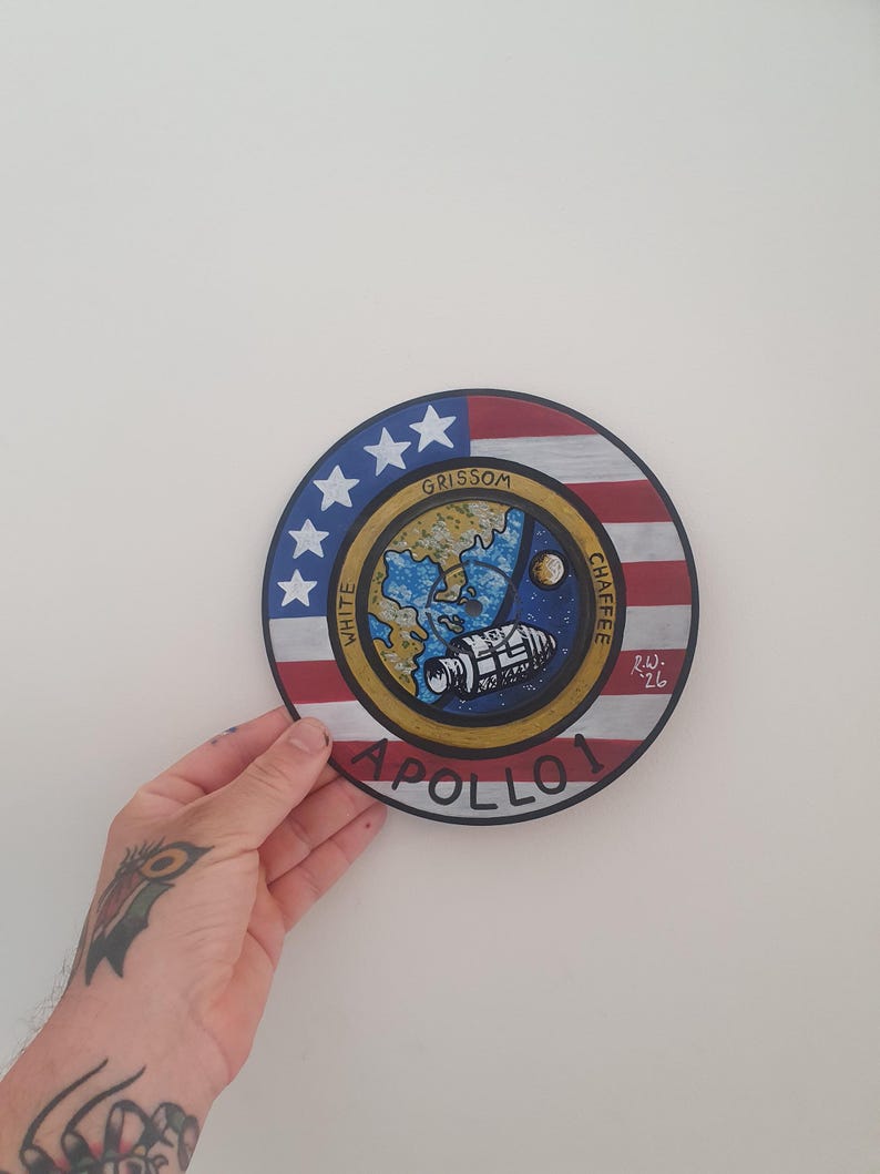 K&ouml;nnte beinhalten: Handgemalte, runde Apollo 1 Gedenktafel. Das Design zeigt ein zentrales Bild eines Raumschiffs, der Erde und des Mondes, umgeben von einem blauen und goldenen Rand. Der &auml;u&szlig;ere Ring zeigt die amerikanische Flagge und die Namen der Astronauten.