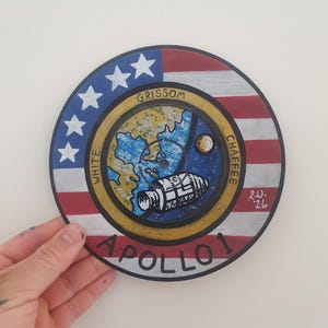 Pode incluir: Uma obra de arte comemorativa do Apollo 1 pintada à mão, de forma redonda. O design apresenta um emblema central com uma cena espacial, rodeado por um anel com os nomes "Grissom", "White" e "Chaffee", e o texto "Apollo 1". O anel exterior tem o design da bandeira americana.