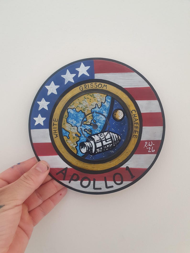 K&ouml;nnte beinhalten: Ein handgemaltes, rundes Apollo 1 Gedenkkunstwerk. Das Design zeigt ein zentrales Emblem mit einer Weltraumszene, umgeben von einem Ring mit den Namen "Grissom", "White" und "Chaffee" sowie dem Text "Apollo 1". Der &auml;u&szlig;ere Ring hat das Design der amerikanischen Flagge.