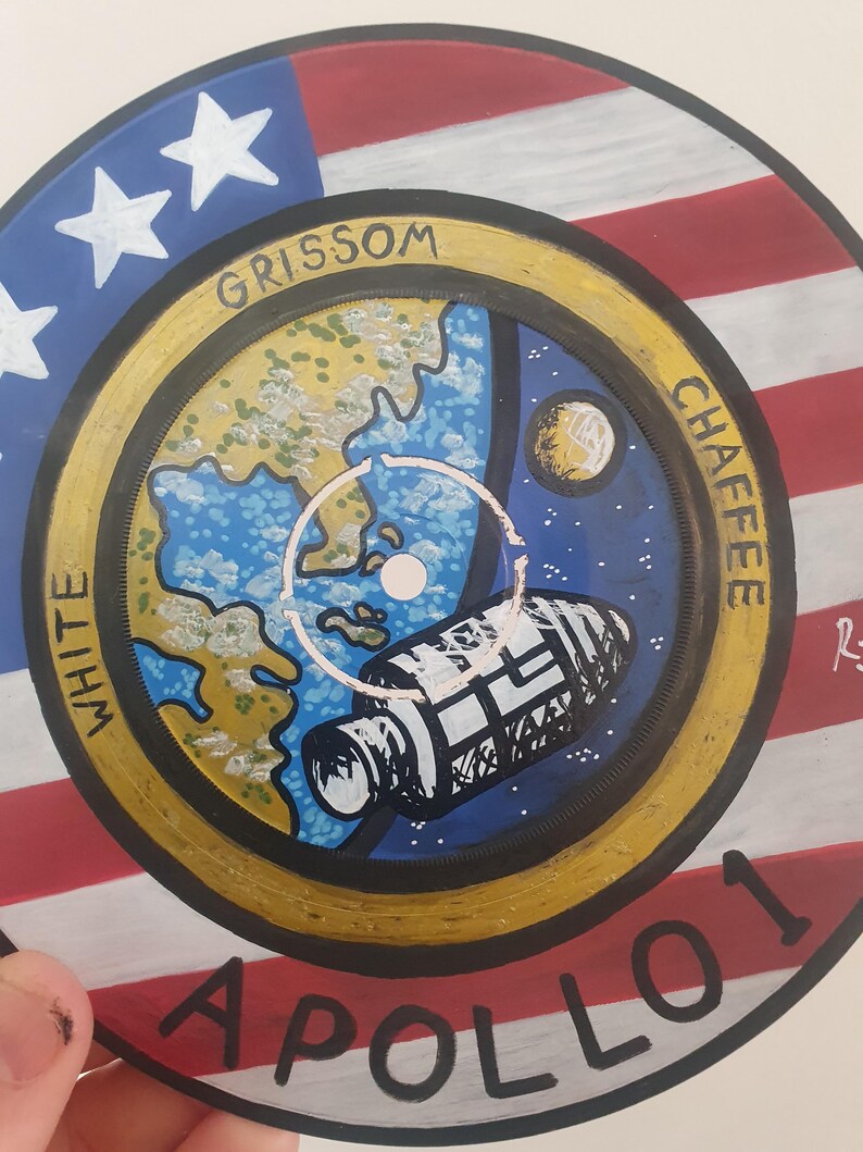 K&ouml;nnte beinhalten: Handbemalte Schallplatte mit einem Apollo 1 Missionsdesign. Das Kunstwerk zeigt einen Hintergrund mit der amerikanischen Flagge, eine Darstellung der Erde, ein Raumschiff und die Namen "Grissom", "Chaffee" und "White". Die Schallplatte ist ein einzigartiges Kunstwerk zum Thema Weltraum.