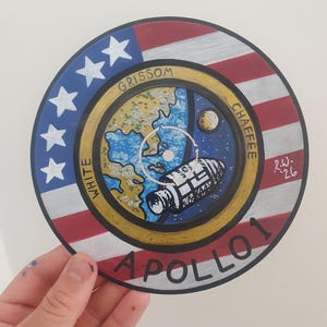 K&ouml;nnte beinhalten: Handgemaltes kreisf&ouml;rmiges Kunstwerk mit dem Emblem der Apollo 1-Mission. Das Design beinhaltet die amerikanische Flagge, Sterne und eine Darstellung der Erde, des Mondes und eines Raumschiffs. Die Namen "Grissom", "White" und "Chaffee" sind sichtbar.