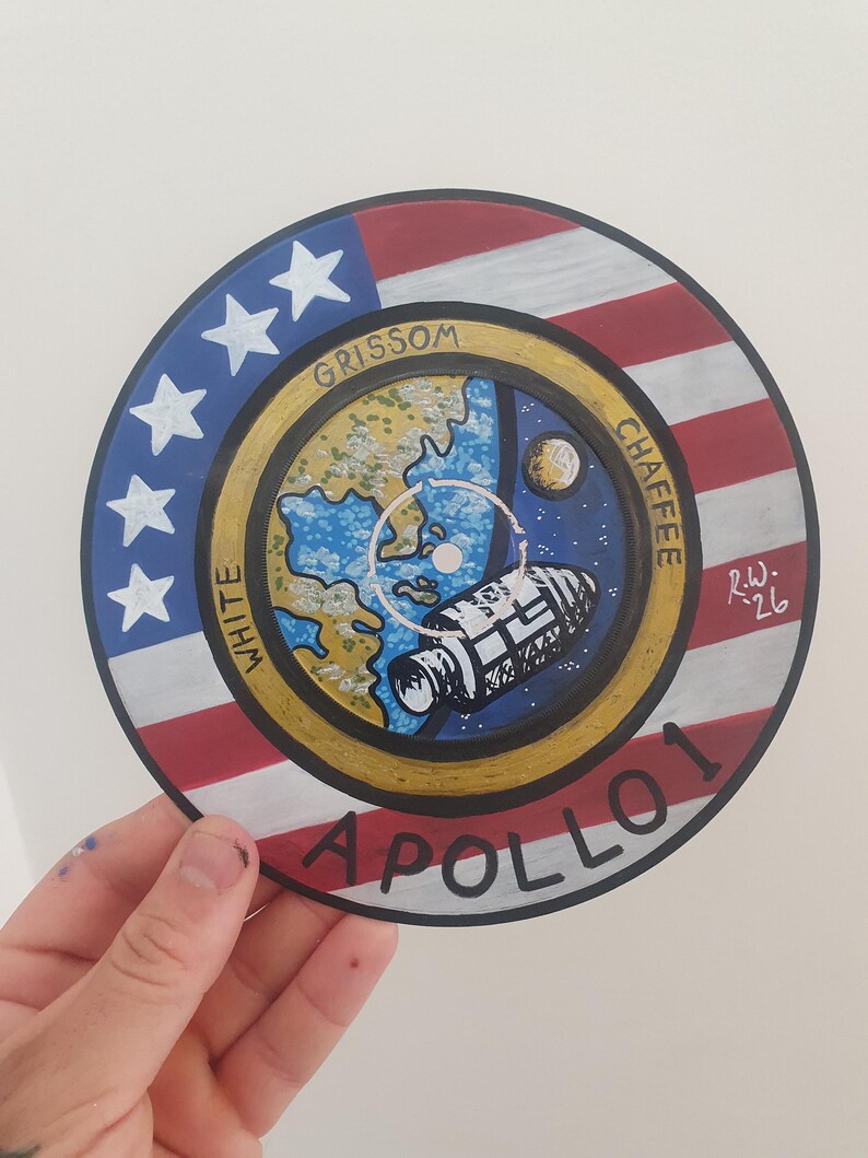K&ouml;nnte beinhalten: Handgemaltes kreisf&ouml;rmiges Kunstwerk mit dem Emblem der Apollo 1-Mission. Das Design beinhaltet die amerikanische Flagge, Sterne und eine Darstellung der Erde, des Mondes und eines Raumschiffs. Die Namen "Grissom", "White" und "Chaffee" sind sichtbar.