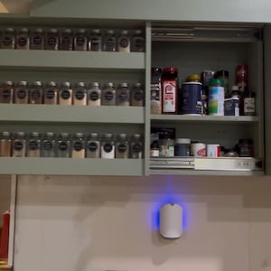 May include: A kitchen spice rack with glass jars. The cabinet is painted a pale green and has a pull-down door. A white air purifier with a blue light is mounted on the wall. The countertop is wooden.