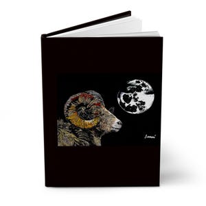 Puede incluir: Un diario de tapa dura negra con una ilustración gráfica de un carnero de cuernos grandes con cuernos curvos y texturizados frente a una luna llena sobre un fondo oscuro. La firma del artista, Curran, es visible en la esquina inferior derecha.