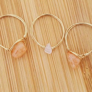 May include: Three rings crafted from thin gold-toned wire, each featuring a single raw gemstone. Two rings are set with irregular, peach-coloured stones, while the centre ring holds a smaller, pale pink, translucent, angular crystal stone.