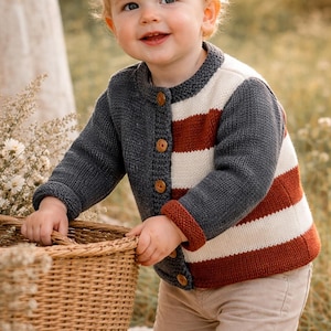 Op de afbeelding: Een kind draagt een gestreept vest in grijs, wit en rood met houten knopen. Het kind houdt een rieten mand vast en glimlacht. Het vest heeft een ronde kraag en geribde manchetten.