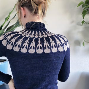 Puede incluir: Jersey de cuello alto azul marino con un patrón geométrico beige en los hombros. El jersey tiene mangas largas y un dobladillo acanalado. La modelo lleva pantalones negros. El jersey está hecho de un material de punto suave.