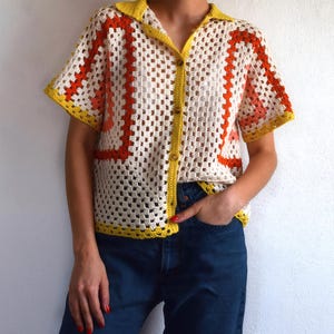 May include: A short-sleeved, button-down crochet shirt. The shirt features a cream-colored base with orange and yellow geometric patterns. The collar and edges are trimmed in yellow. The shirt is worn with blue jeans.