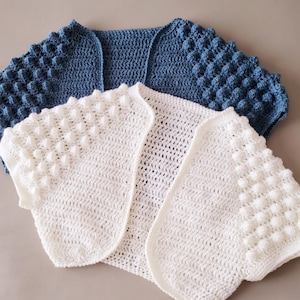 May include: Two handmade crochet shrugs. One is a solid blue, the other is white. Both have a textured, bobble stitch pattern on the sleeves. The white shrug has a delicate open weave. The shrugs are displayed on a neutral background.