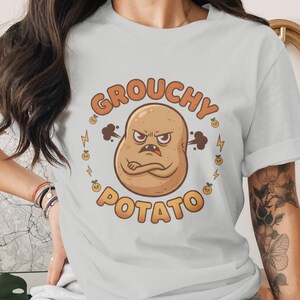 May include: Light blue t-shirt featuring a cartoon illustration of a grumpy potato with crossed arms. The text "GROUCHY POTATO" is in orange, encircling the potato. The design includes lightning bolts and small orange potato faces.