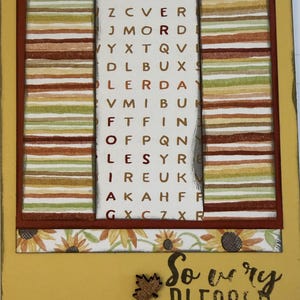 May include: A rectangular greeting card with a yellow border featuring a central panel of a word search puzzle with the word FOLIAGE highlighted in red. The card includes side panels with horizontal stripes in green, orange, and brown, a sunflower border, and a small wooden maple leaf cutout next to the text So very BLESSED.