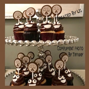 May include: A two-tiered silver cake stand with cupcakes decorated with chocolate frosting and googly eyes. Each cupcake has a brown and white circular topper with the text "Holy Cow! Digger 40".