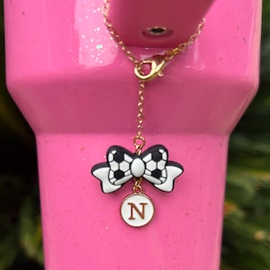 May include: A gold-colored chain with a black and white soccer ball bow charm and a white circular charm with the letter "N" on a pink tumbler. The charm is attached to the tumbler's lid.