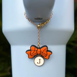 May include: A gold-colored chain with a lobster clasp is attached to a light blue tumbler. A charm hangs from the chain, featuring an orange and black bow design and a white circular pendant with the letter "J".