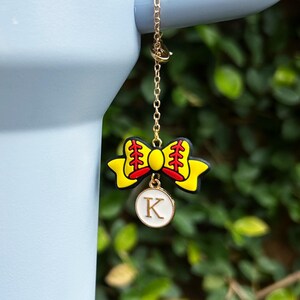 May include: A gold-colored chain with a yellow bow-shaped charm featuring red and yellow details. A white circular charm with the letter "K" is suspended below the bow. The background is a blurred green.